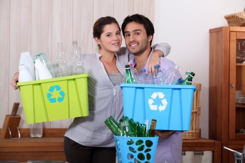 A young couple stands indoors in a room with light-colored wooden paneling, holding three plastic recycling bins filled with empties. The woman, on the left, holds a bright green bin with a recycling symbol on the front, containing clear glass bottles. She has dark hair pulled back, a light gray hoodie, and a neutral expression. The man, on the right, holds a blue bin with a recycling symbol, filled with clear plastic bottles, and is wearing a plaid shirt over a T-shirt, smiling at the camera. Between them, a smaller blue container holds additional empty glass bottles. The background shows a wooden sideboard with woven baskets and decorative items, indicating a residential setting. The image emphasizes proper waste segregation and recycling practice, relevant to rubbish removal services provided by Commercial Waste Finsbury Park in the local area.