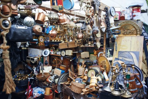 The image displays a densely packed outdoor market stall or pile of various discarded household items and vintage objects, including numerous copper and brass kitchenware pieces such as pots, pans, kettles, and decorative trays, all with a aged and tarnished finish. There are also ceramic and glass items, some partly broken or chipped, along with small wooden and wicker objects scattered across the cluttered surface. In the background, overlapping shelves and the surrounding clutter emphasize an overflowing display, with some objects leaning against each other or stacked haphazardly. The environment suggests an outdoor setting, possibly a market or junkyard, with ambient natural light illuminating the diverse textures and muted colors of the mixed materials. The scene is consistent with rubbish or waste collection, indicative of items awaiting disposal or recycling, which aligns with services offered by Commercial Waste Finsbury Park in the vicinity of N4 postal codes. The overall impression is of a large, chaotic assortment of used goods and discarded items that require professional rubbish removal or clearance services.