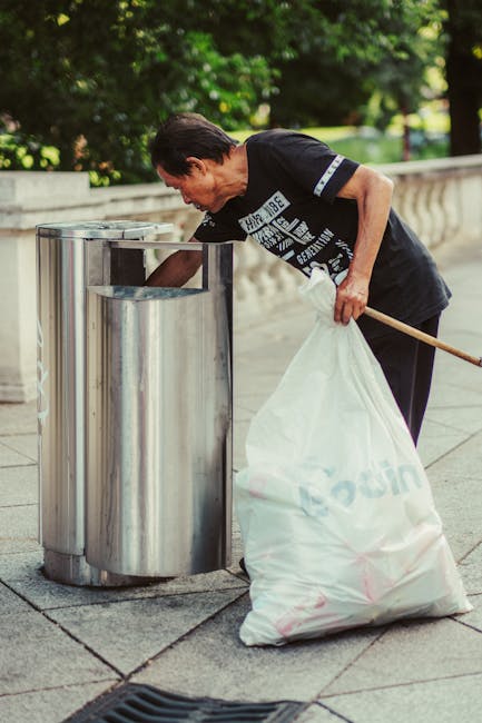 A green outdoor public waste bin with a rounded top and transparent section revealing various rubbish, including paper and plastic items. The bin has a blue plastic bag lining the interior and a black