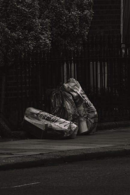 Two large black plastic garbage bags filled with mixed waste sit on a concrete pavement in front of a black metal fence and dark shrubbery. The bags are crumpled and appear to contain domestic rubbish
